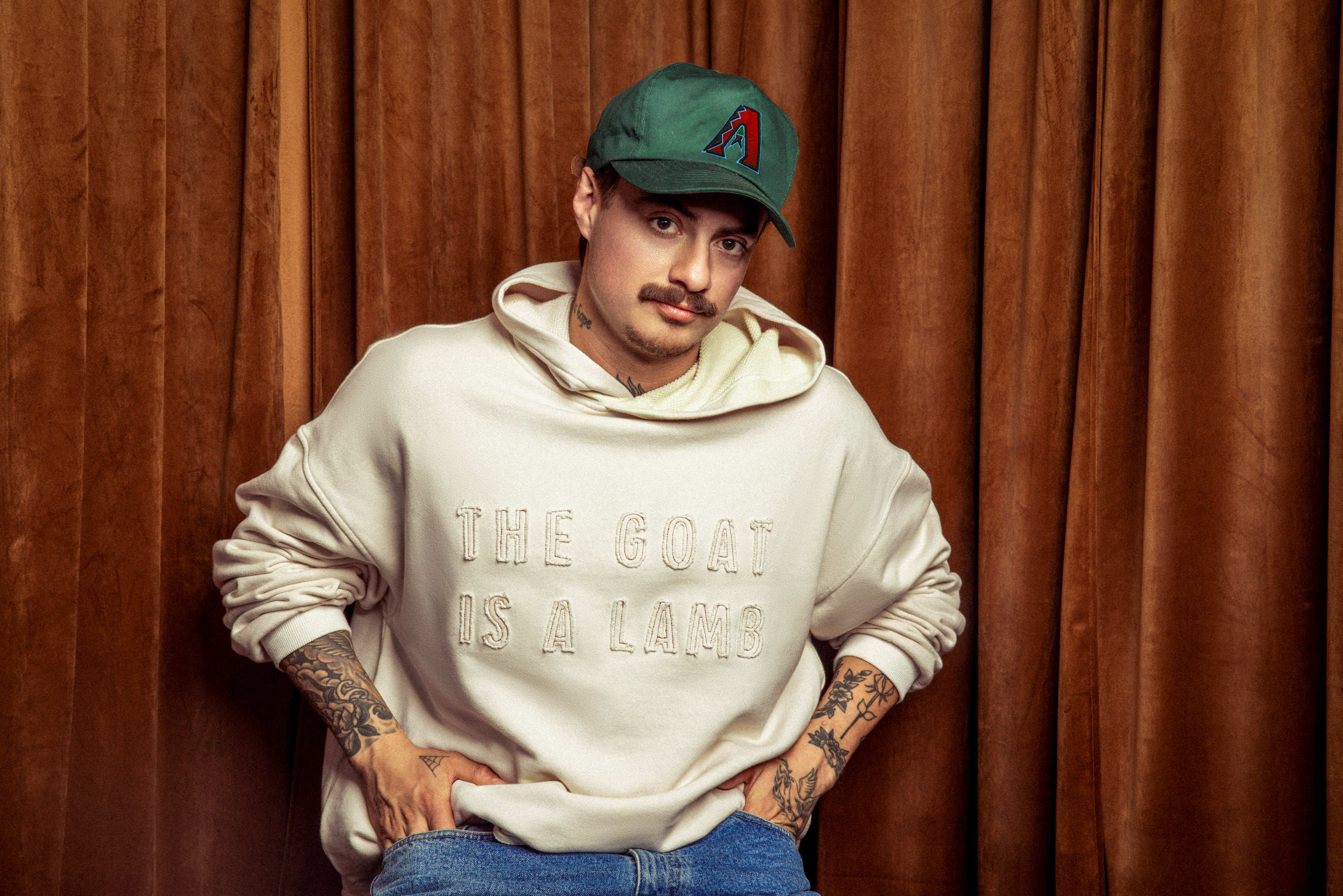 Person wearing a beige The Goat Is A Lamb hoodie and a green cap, sitting on a stool against a brown curtain.